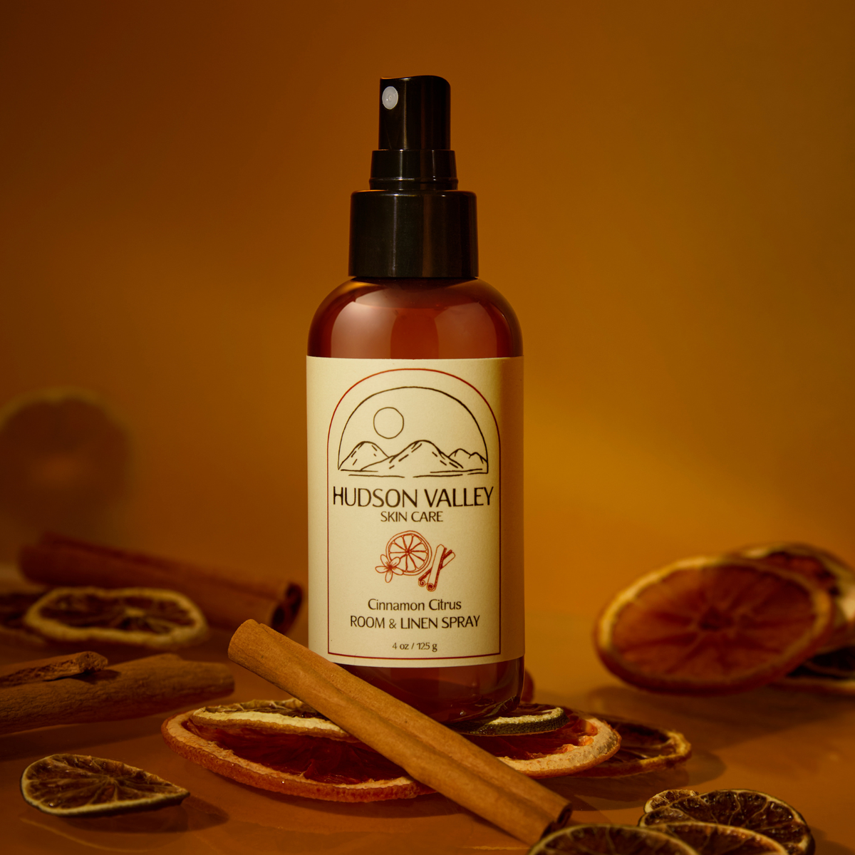 A bottle of Hudson Valley Skin Care Cinnamon Citrus Room & Linen Spray stands centered against a warm amber background, surrounded by dried orange slices, cinnamon sticks, and a few whole citrus pieces. The bottle is amber glass with a black spray top and a cream label featuring the Hudson Valley logo.