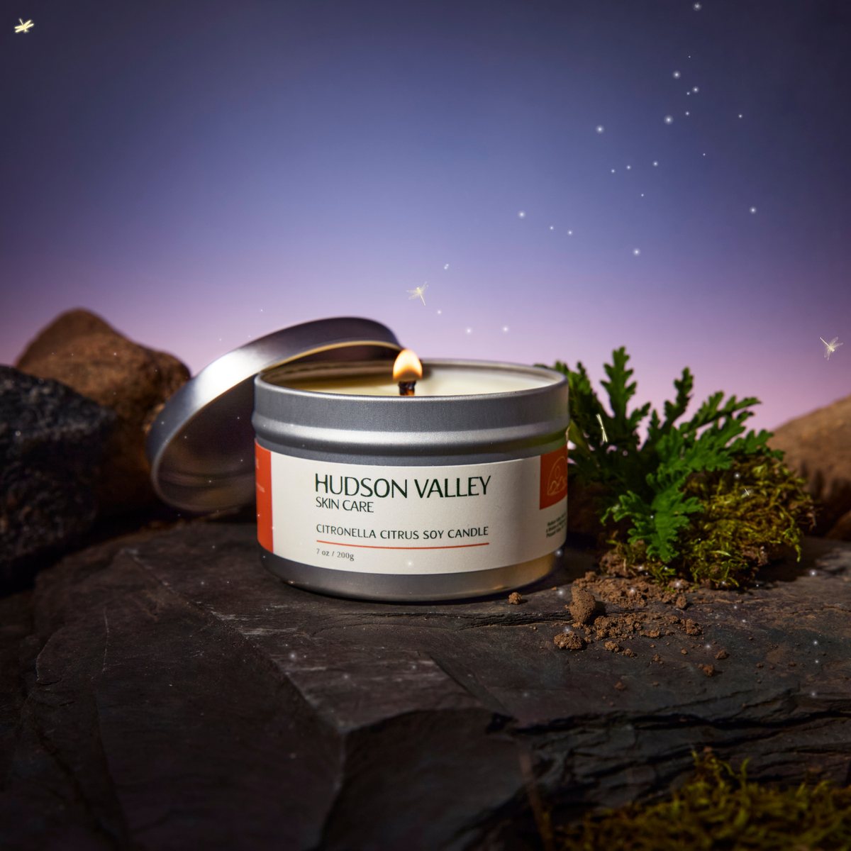 A lit Hudson Valley Skin Care citronella citrus soy candle in a silver tin, styled on a dark slate rock with moss and small rocks against a purple starry sky background.