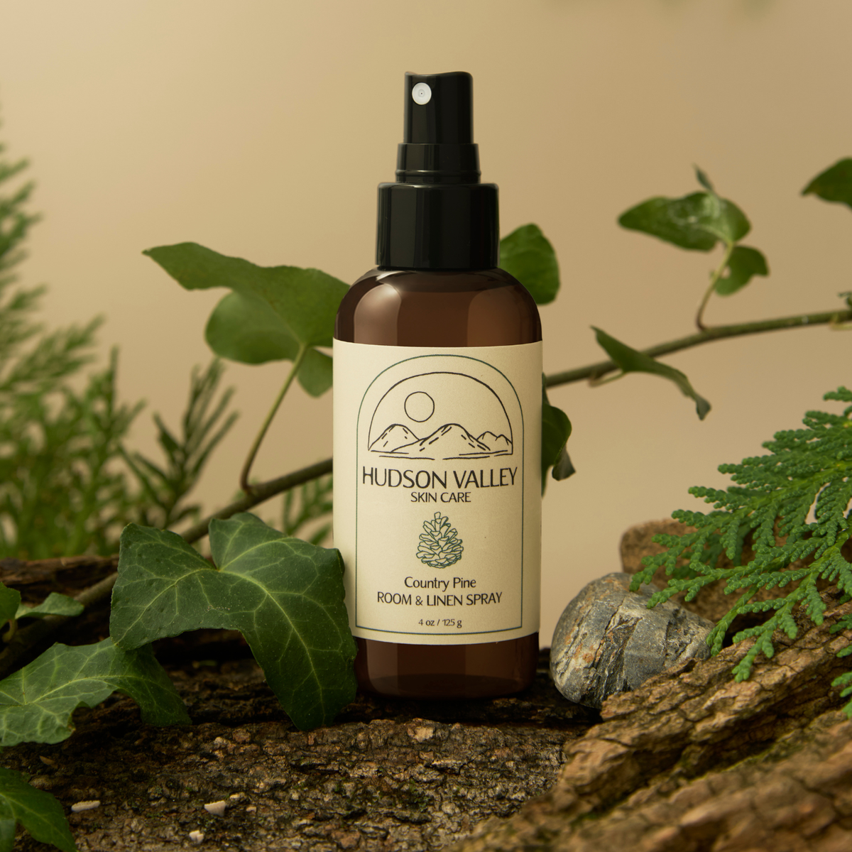 A bottle of Hudson Valley Skin Care 'Country Pine' room and linen spray, styled on a rustic log with green leaves and pine branches against a warm beige background.