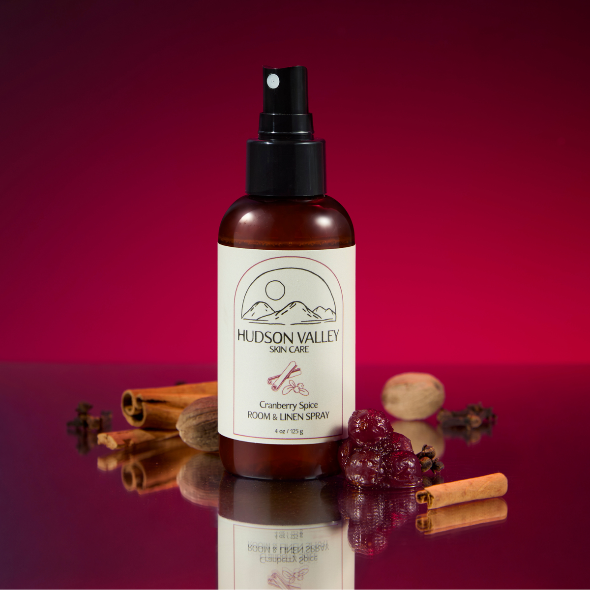 A bottle of Hudson Valley Skin Care Cranberry Spray stands on a reflective surface against a deep red gradient background, surrounded by cranberries, cinnamon sticks, and a nut. The amber bottle has a black spray top and a cream label with the Hudson Valley Skin Care logo.