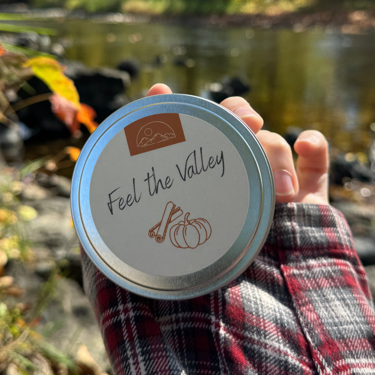 A hand wearing a red and black plaid flannel shirt holds a round silver tin labeled 'Feel the Valley' with a pumpkin on it, set against a blurred outdoor background of a river and autumn trees.