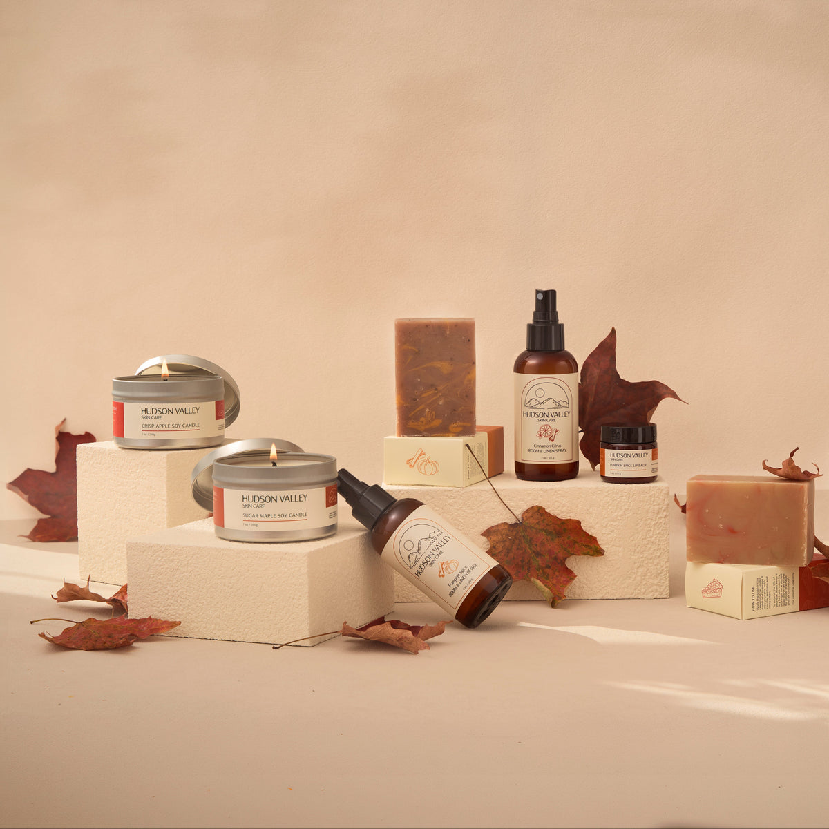 A curated collection of natural skincare and bath products arranged on minimalist pedestals with autumn maple leaves. The products include amber glass bottles, metal tins, a pump bottle, and solid bars of soap in earthy tones, set against a warm beige background.