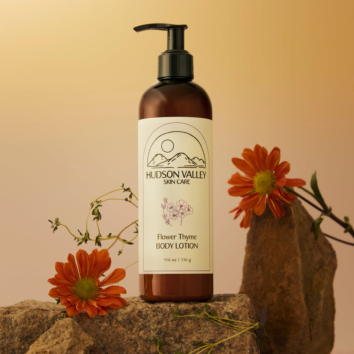A bottle of Hudson Valley Skin Care Flower Thyme Body Lotion stands on a stone prop against a warm beige background, decorated with orange flowers and green stems.