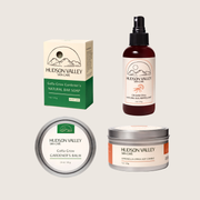 Collection of Hudson Valley Skin Care garden products including a bar soap, balm, candle, and spray bottle on a light beige background.