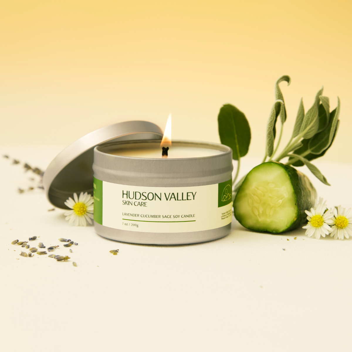 A lit Hudson Valley Skin Care candle in a silver tin with its lid leaning beside it, styled with a cucumber slice, sage leaves, daisies, and scattered lavender on a warm cream surface against a soft yellow background.