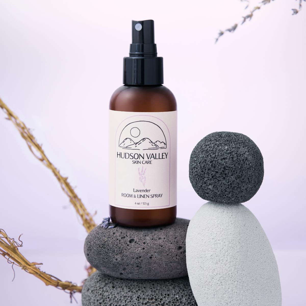 Hudson Valley Skin Care Lavender Room & Linen Spray in an amber bottle with a black spray top, styled on stacked volcanic rocks with dried branches against a soft lavender background.