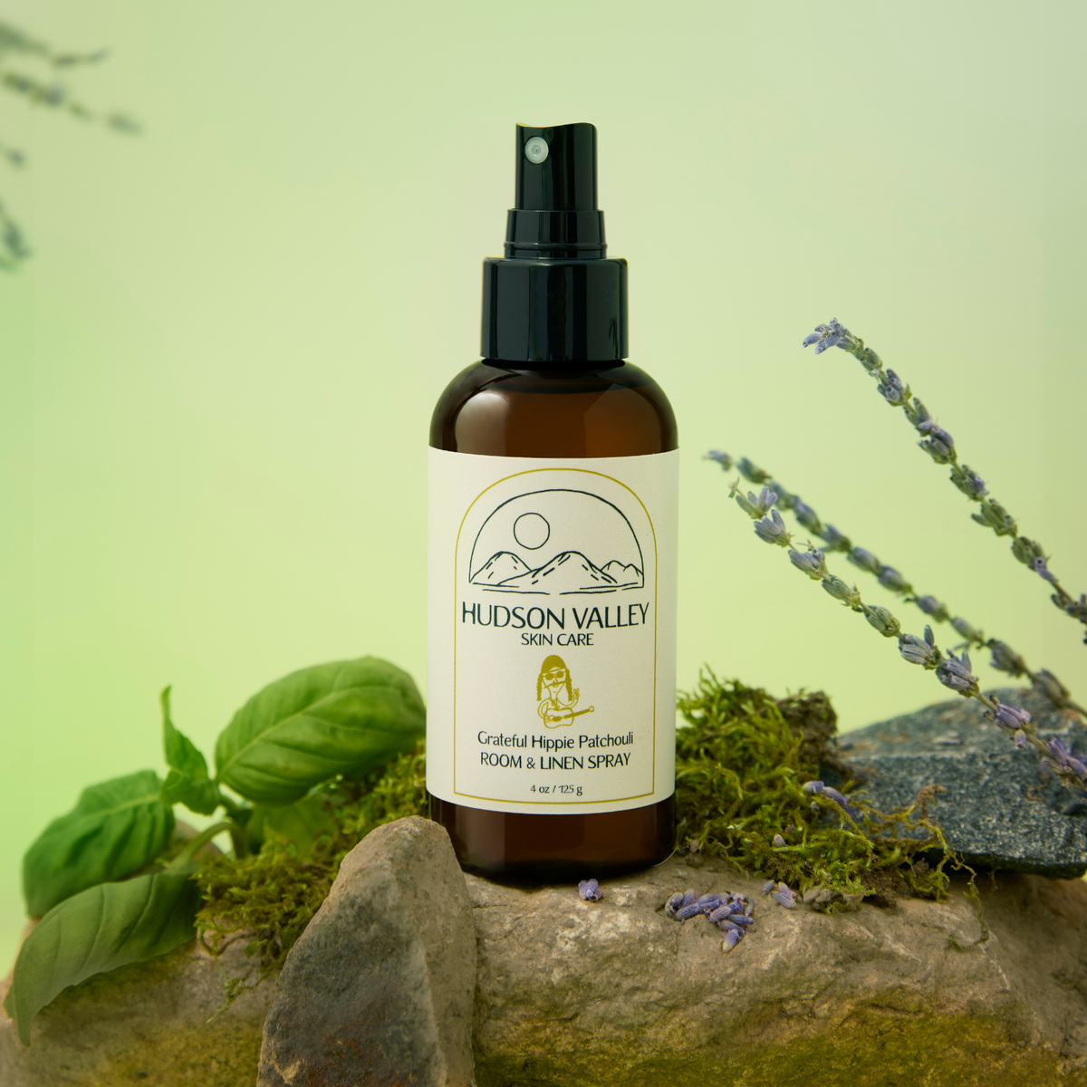 A bottle of Hudson Valley Skin Care Grateful Hippie Patchouli Room & Linen Spray, set on a mossy rock with basil leaves and lavender sprigs against a soft green background.