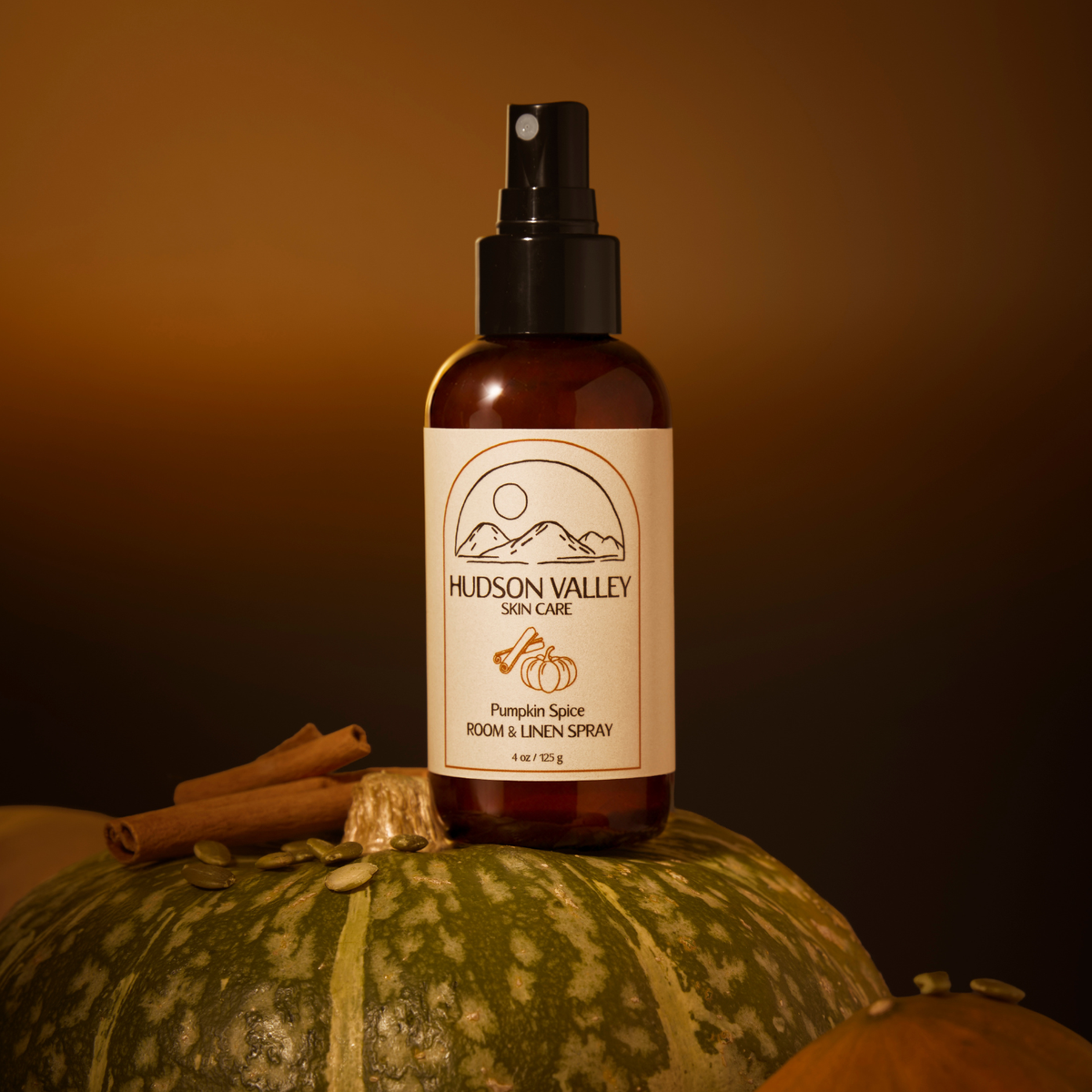 A bottle of Hudson Valley Skin Care Pumpkin Spice Room & Linen Spray stands on a green striped pumpkin with cinnamon sticks against a warm brown background.