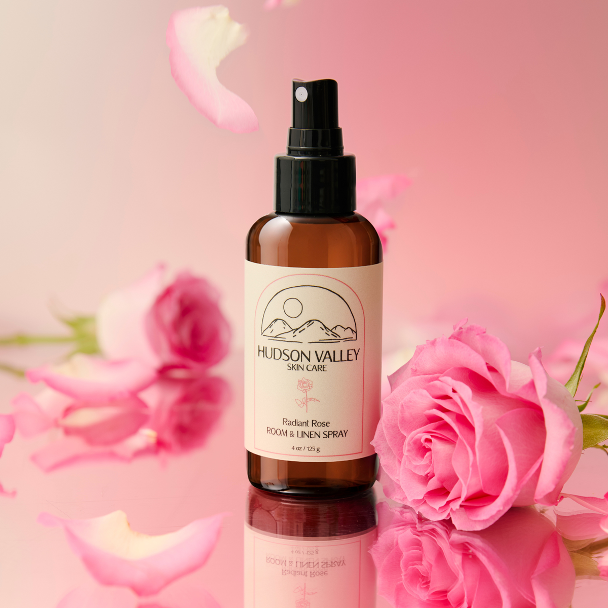 A bottle of Hudson Valley Skin Care Radiant Rose Room & Lipen Spray, set against a soft pink background with pink roses and petals. The amber glass bottle has a black spray pump and a cream-colored label with a mountain logo.