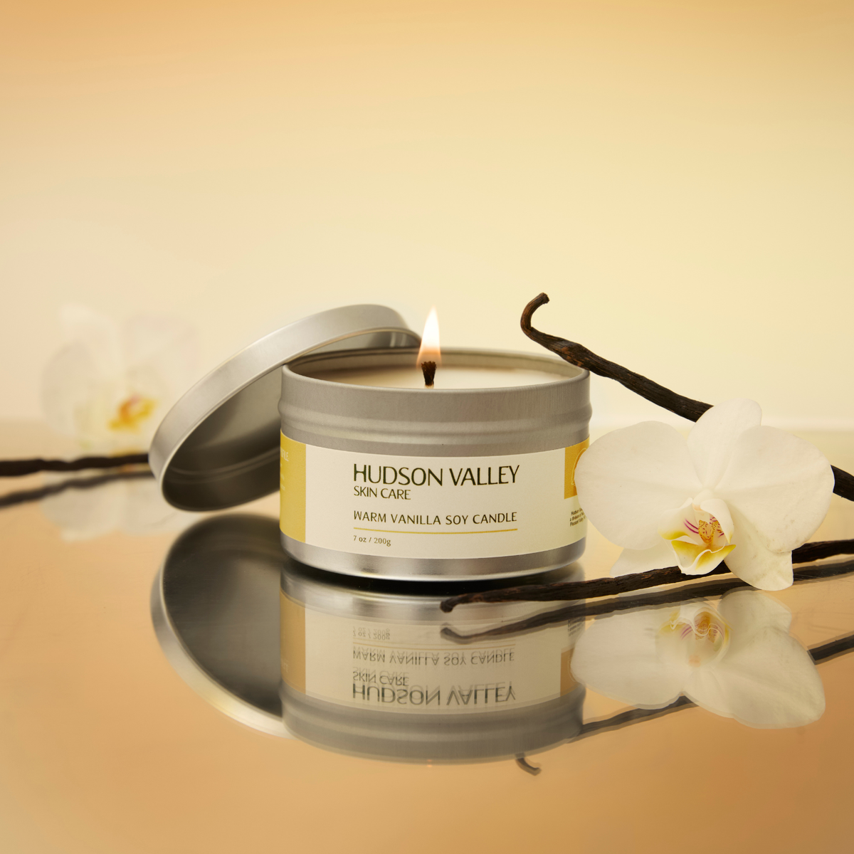 A lit Hudson Valley Skin Care 'Warm Vanilla Soy Candle' in a silver tin with a white label, styled with a vanilla bean and white orchids on a reflective beige surface.